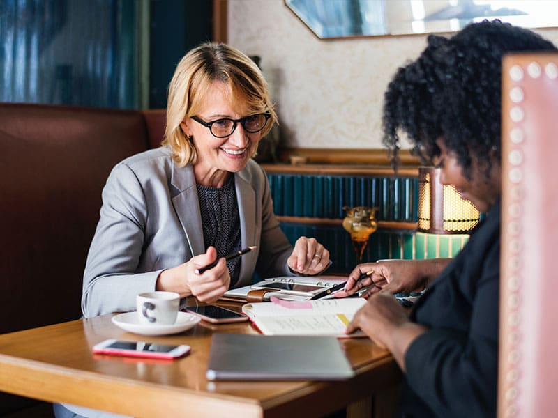 Two women discussing work at a cafe table with notebooks and coffee.