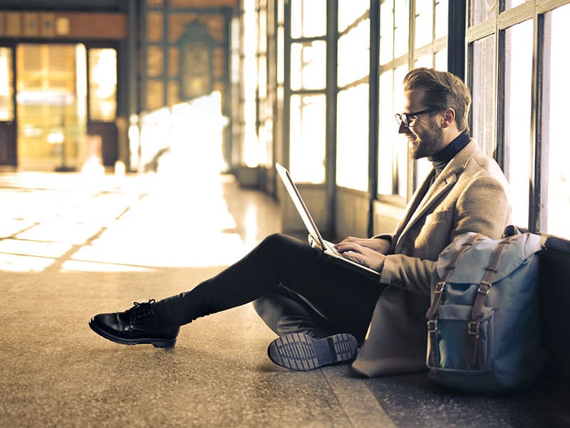 Man in beige coat working on laptop while sitting on floor near windows.