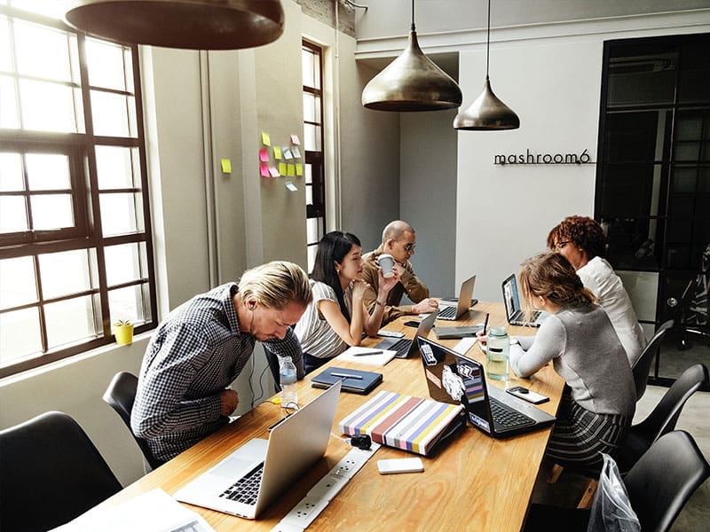 A diverse group collaborating around a table with laptops in a modern meeting room.