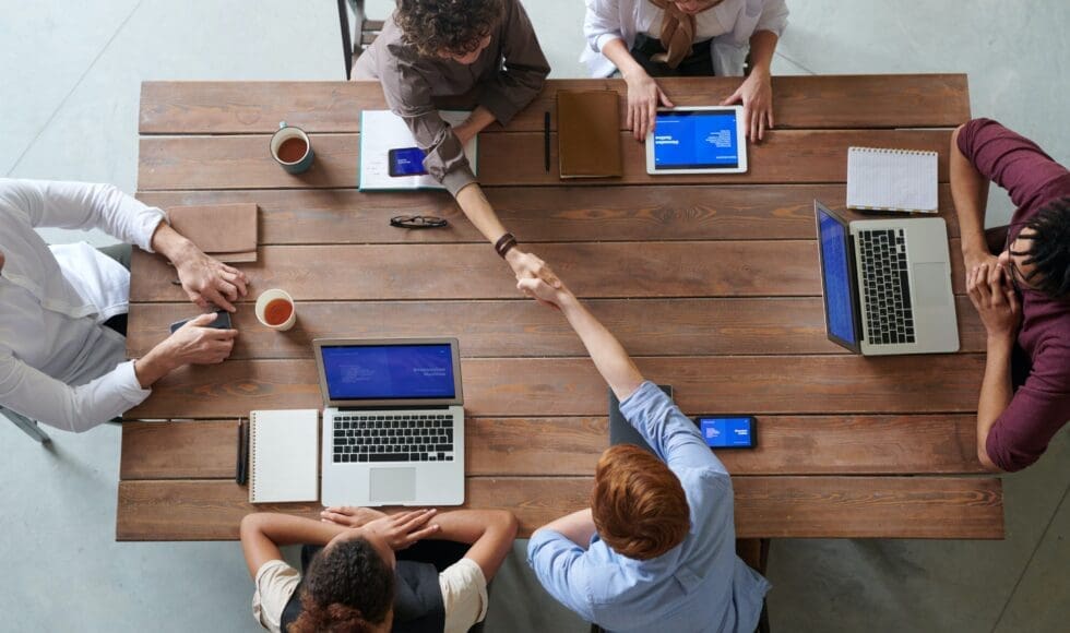 A group of people collaborating around a wooden table with laptops and tablets.