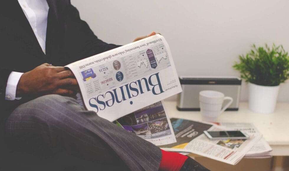 Person reading a business newspaper at a table with coffee and a laptop.