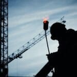 Silhouette of a construction worker with a crane and torch flame in the background.
