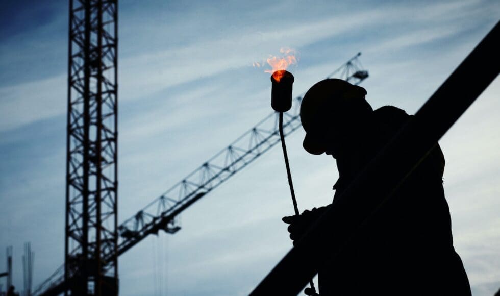 Silhouette of a construction worker with a crane and torch flame in the background.