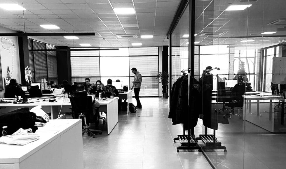 Modern office with employees working and a person walking near large windows.