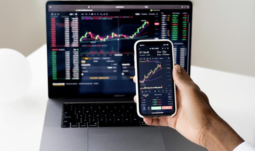 Person viewing cryptocurrency charts on phone and laptop screens.